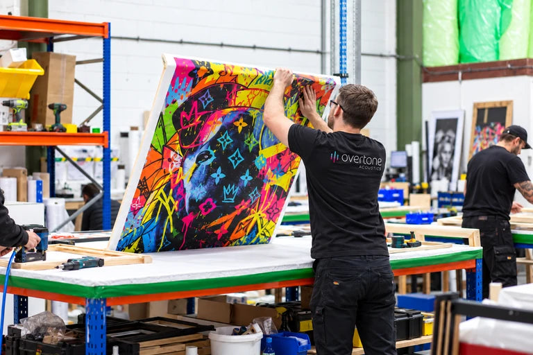 Illustrative workshop scene of a technician wrapping colorful printed canvas around a panel frame.